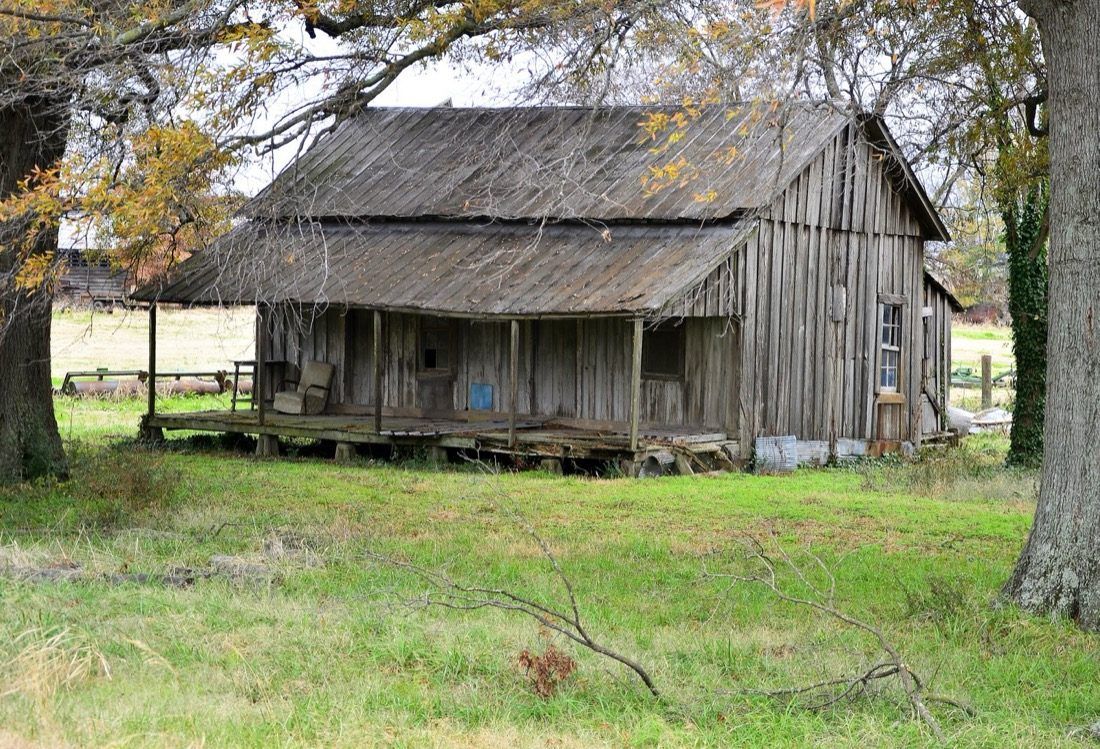 An abandoned single-story home in rural Tunica County, Mississippi, with a sagging roofline and weathered wood siding against an overcast sky. Contemporary photograph of housing stock in the county that ranks third of 3,144 nationally for household financial distress.