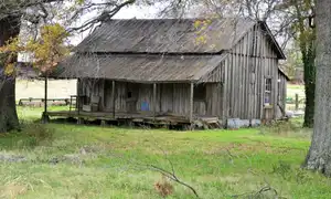 An abandoned single-story home in rural Tunica County, Mississippi, with a sagging roofline and weathered wood siding against an overcast sky. Contemporary photograph of housing stock in the county that ranks 10th of 3,144 nationally for household financial distress.